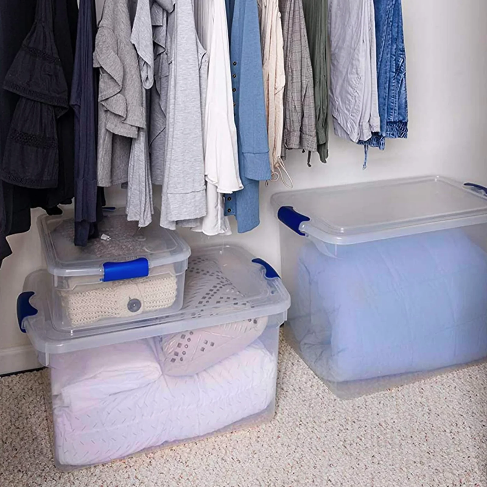 Clear HOMZ Secure Latch bins of different sizes on a closet floor under hanging clothes, a small bin stacked on a wider one and a tall bin beside them.