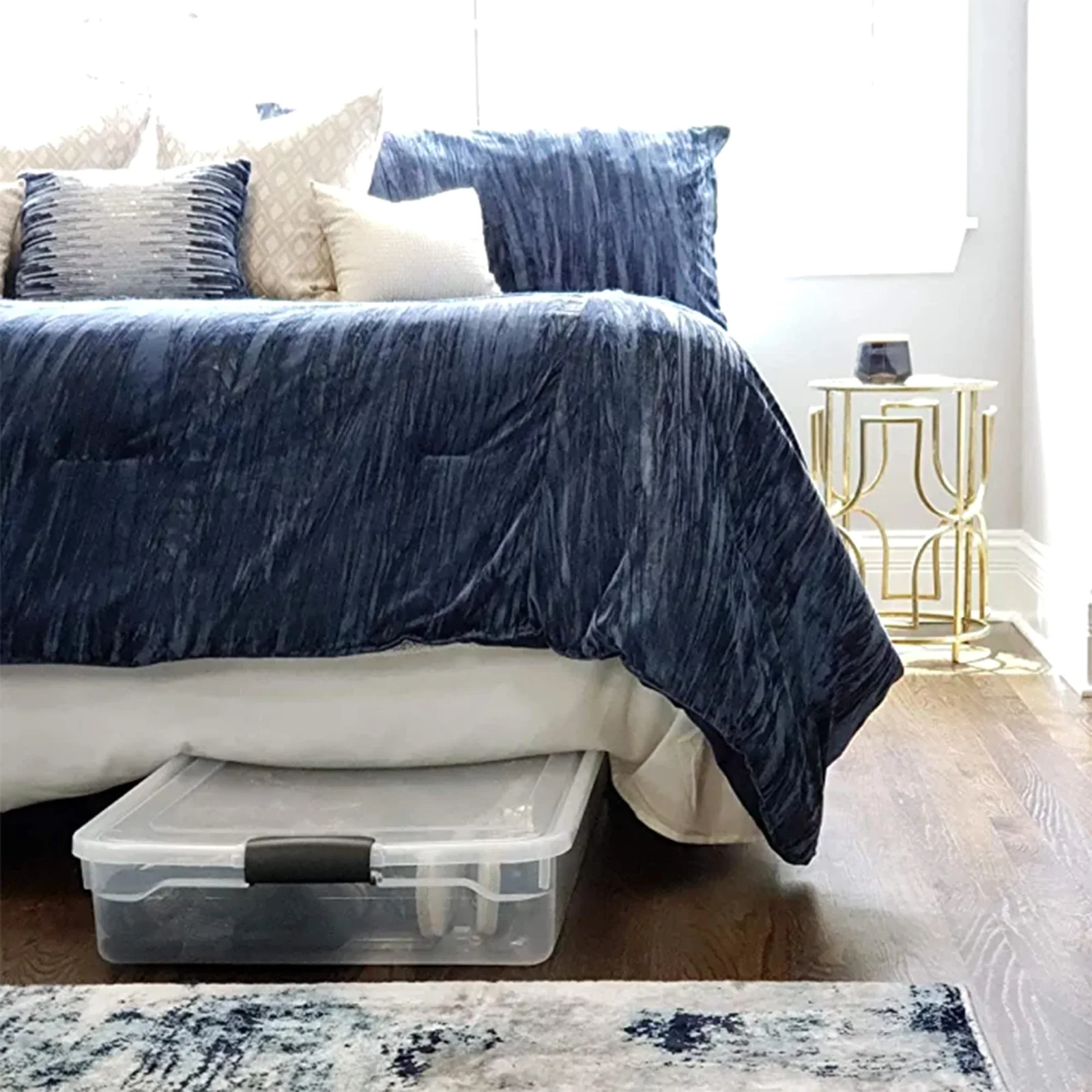 A clear HOMZ 60-quart underbed bin with dark latches, pulled halfway out from under a bed with a blue comforter.