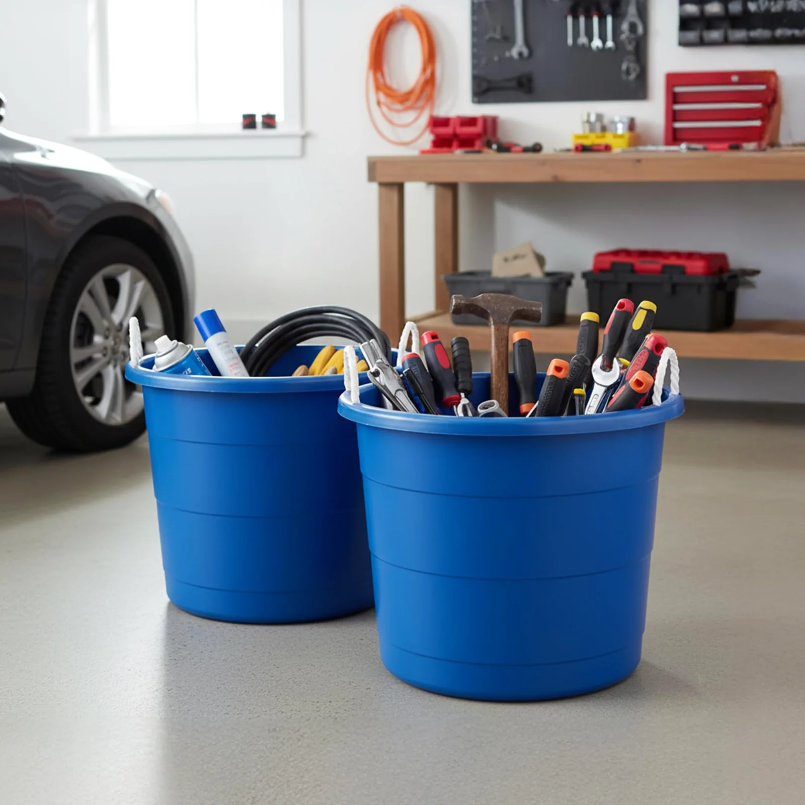 Two blue HOMZ rope-handle tubs on a garage floor, one packed with screwdrivers and a hammer, with a workbench and a pegboard of wrenches behind.