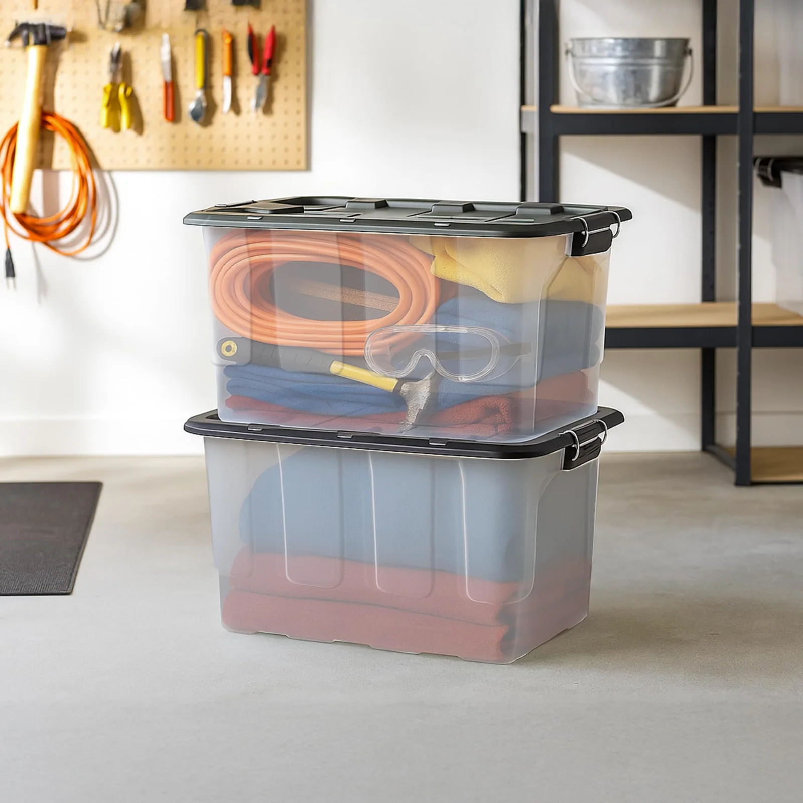 Two clear HOMZ 30-gallon totes with black lids stacked in a garage, orange extension cords and safety glasses showing through the top one, a pegboard of tools behind.